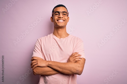 Handsome african american man wearing casual t-shirt and glasses over pink background happy face smiling with crossed arms looking at the camera. Positive person. © Krakenimages.com
