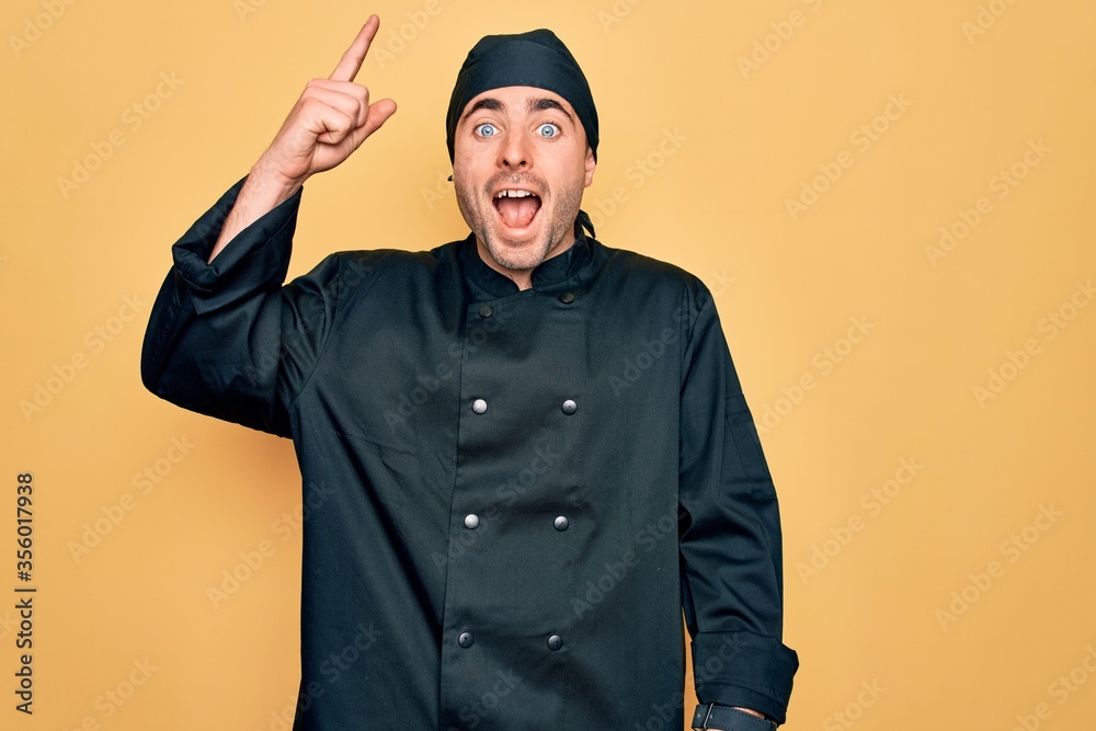 Young handsome cooker man with blue eyes wearing uniform and hat over yellow background pointing finger up with successful idea. Exited and happy. Number one. Young handsome cooker man with blue eyes wearing uniform and hat over yellow background pointing finger up with successful idea. Exited and happy. Number one.