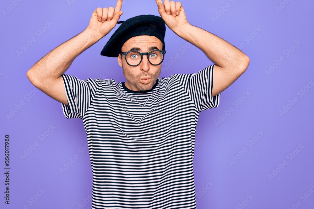 Naklejka premium Handsome man with blue eyes wearing striped t-shirt and french beret over purple background doing funny gesture with finger over head as bull horns