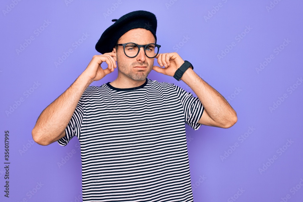 Handsome man with blue eyes wearing striped t-shirt and french beret over purple background covering ears with fingers with annoyed expression for the noise of loud music. Deaf concept. Handsome man with blue eyes wearing striped t-shirt and french beret over purple background covering ears with fingers with annoyed expression for the noise of loud music. Deaf concept.