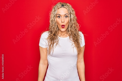 Young beautiful blonde woman wearing casual t-shirt standing over isolated red background afraid and shocked with surprise expression, fear and excited face.