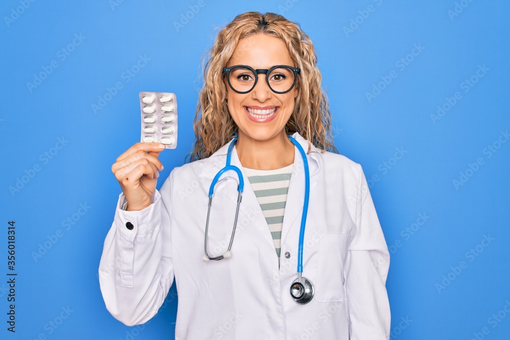 Young beautiful blonde cardiologist woman wearing stethoscope holding plastic heart looking positive and happy standing and smiling with a confident smile showing teeth