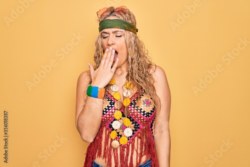 Beautiful blonde hippie woman wearing sunglasses and accessories over yellow background bored yawning tired covering mouth with hand. Restless and sleepiness.