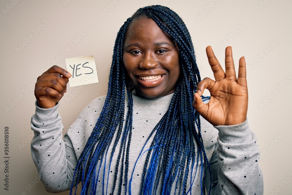 Young african american plus size woman with braids holding reminder paper with yes message doing ok sign with fingers, excellent symbol Young african american plus size woman with braids holding reminder paper with yes message doing ok sign with fingers, excellent symbol