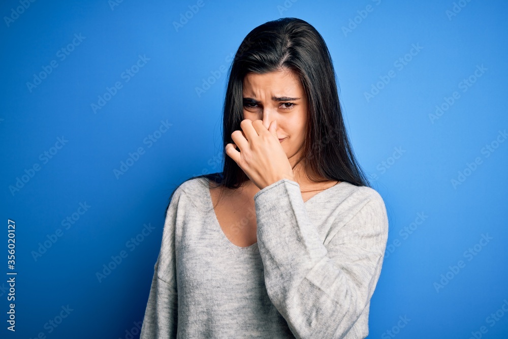 Young beautiful brunette woman wearing casual sweater standing over blue background smelling something stinky and disgusting, intolerable smell, holding breath with fingers on nose. Bad smell Young beautiful brunette woman wearing casual sweater standing over blue background smelling something stinky and disgusting, intolerable smell, holding breath with fingers on nose. Bad smell