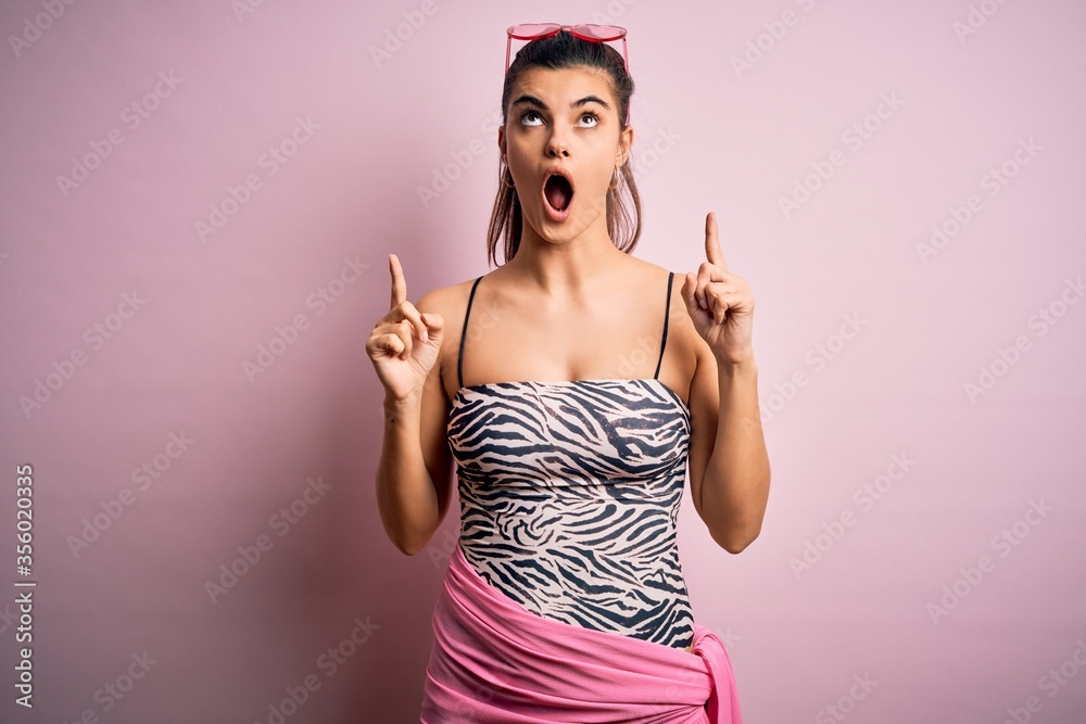 Φωτογραφία Young beautiful brunette woman on vacation wearing swimsuit over pink background amazed and surprised looking up and pointing with fingers and raised arms