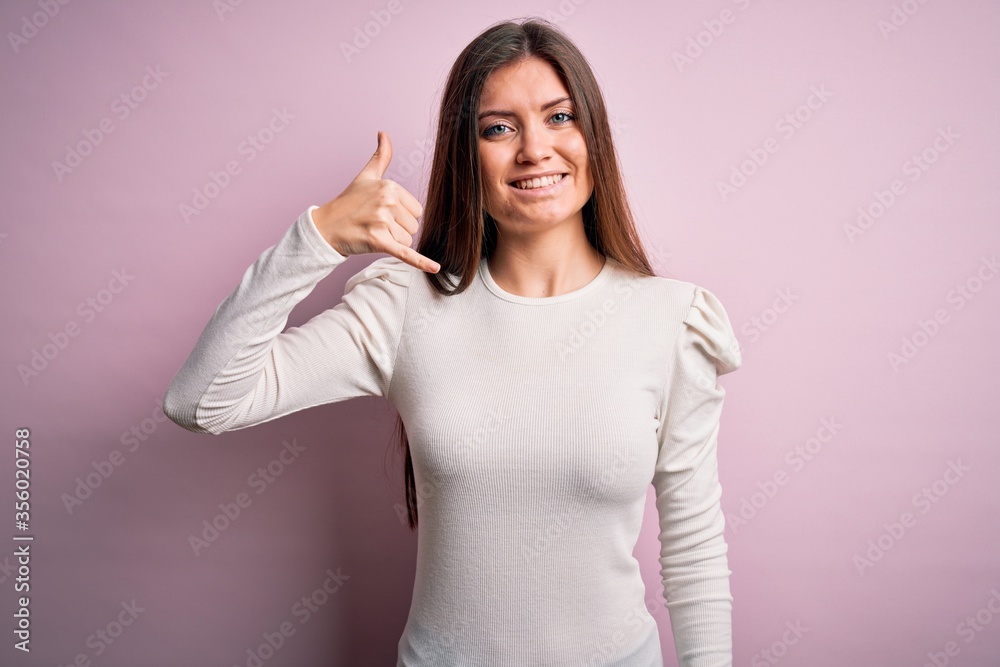 Young beautiful woman with blue eyes wearing casual white t-shirt over pink background smiling doing phone gesture with hand and fingers like talking on the telephone. Communicating concepts. Young beautiful woman with blue eyes wearing casual white t-shirt over pink background smiling doing phone gesture with hand and fingers like talking on the telephone. Communicating concepts.