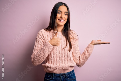 Young beautiful brunette woman wearing casual sweater over isolated pink background Showing palm hand and doing ok gesture with thumbs up, smiling happy and cheerful