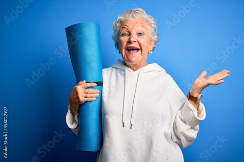 Senior beautiful sporty woman holding mat for yoga standing over isolated blue background very happy and excited, winner expression celebrating victory screaming with big smile and raised hands