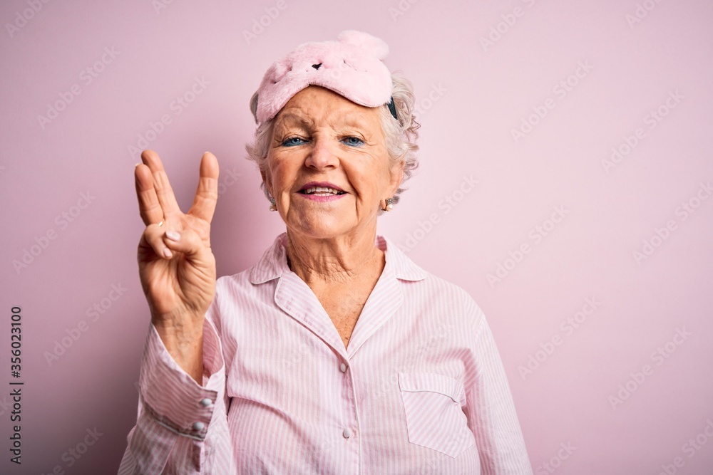 Senior beautiful woman wearing sleep mask and pajama over isolated pink background showing and pointing up with fingers number three while smiling confident and happy.