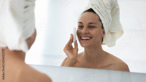 Close up head shot smiling woman cleaning skin with facial cleansing sponge, attractive girl wearing white towel on head looking in mirror, enjoying face care procedure, removing makeup