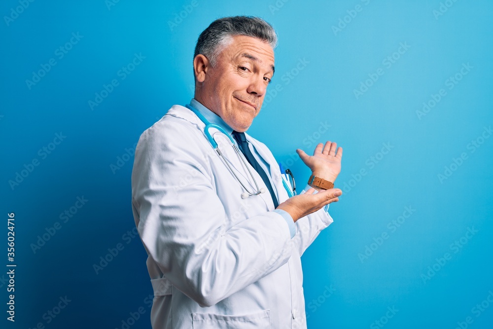 Middle age handsome grey-haired doctor man wearing coat and blue stethoscope Inviting to enter smiling natural with open hand Middle age handsome grey-haired doctor man wearing coat and blue stethoscope Inviting to enter smiling natural with open hand