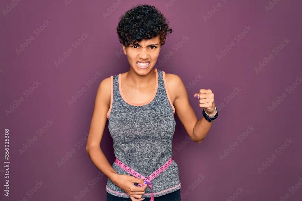 Beautiful african american sporty woman doing exercise controlling weight using tape measure annoyed and frustrated shouting with anger, crazy and yelling with raised hand, anger concept