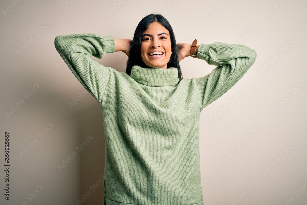 Young beautiful hispanic woman wearing green winter sweater over isolated background relaxing and stretching, arms and hands behind head and neck smiling happy Young beautiful hispanic woman wearing green winter sweater over isolated background relaxing and stretching, arms and hands behind head and neck smiling happy