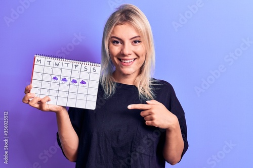 Beautiful blonde woman holding weather calendar showing rainy week over purple background smiling happy pointing with hand and finger