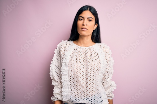 Young beautiful hispanic fashion woman wearing elegant shirt over pink background with serious expression on face. Simple and natural looking at the camera.