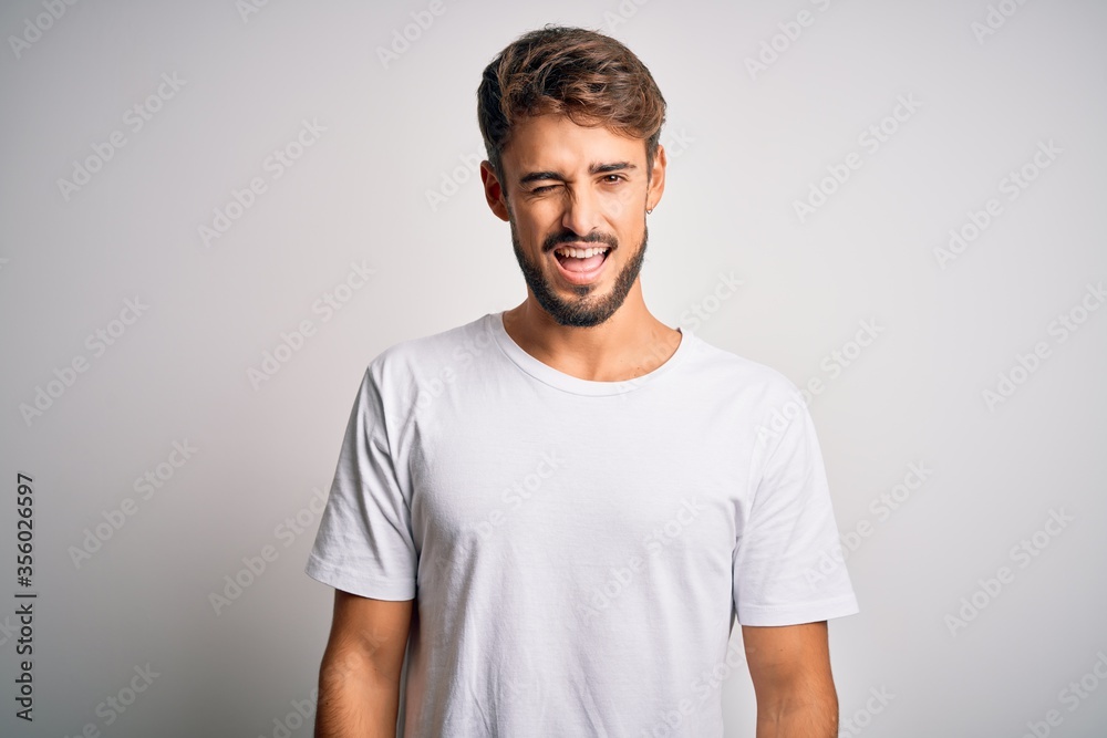 Young handsome man with beard wearing casual t-shirt standing over white background winking looking at the camera with sexy expression, cheerful and happy face. Young handsome man with beard wearing casual t-shirt standing over white background winking looking at the camera with sexy expression, cheerful and happy face.