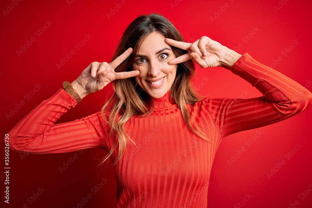 Young beautiful brunette woman wearing casual turtleneck sweater over red background Doing peace symbol with fingers over face, smiling cheerful showing victory