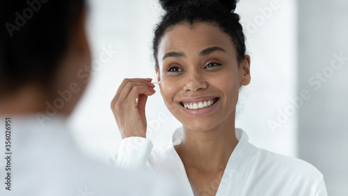 Mirror reflection head shot close up smiling African American young woman cleaning ears, beautiful girl wearing white bathrobe using cotton bud after shower, morning routine, personal hygiene