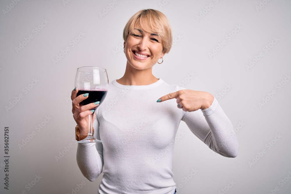 Young blonde woman with short hair drinking a glass of red wine over isolated background with surprise face pointing finger to himself Young blonde woman with short hair drinking a glass of red wine over isolated background with surprise face pointing finger to himself
