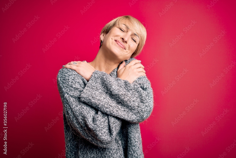 Young blonde woman with modern short hair wearing casual sweater over pink background Hugging oneself happy and positive, smiling confident. Self love and self care Young blonde woman with modern short hair wearing casual sweater over pink background Hugging oneself happy and positive, smiling confident. Self love and self care