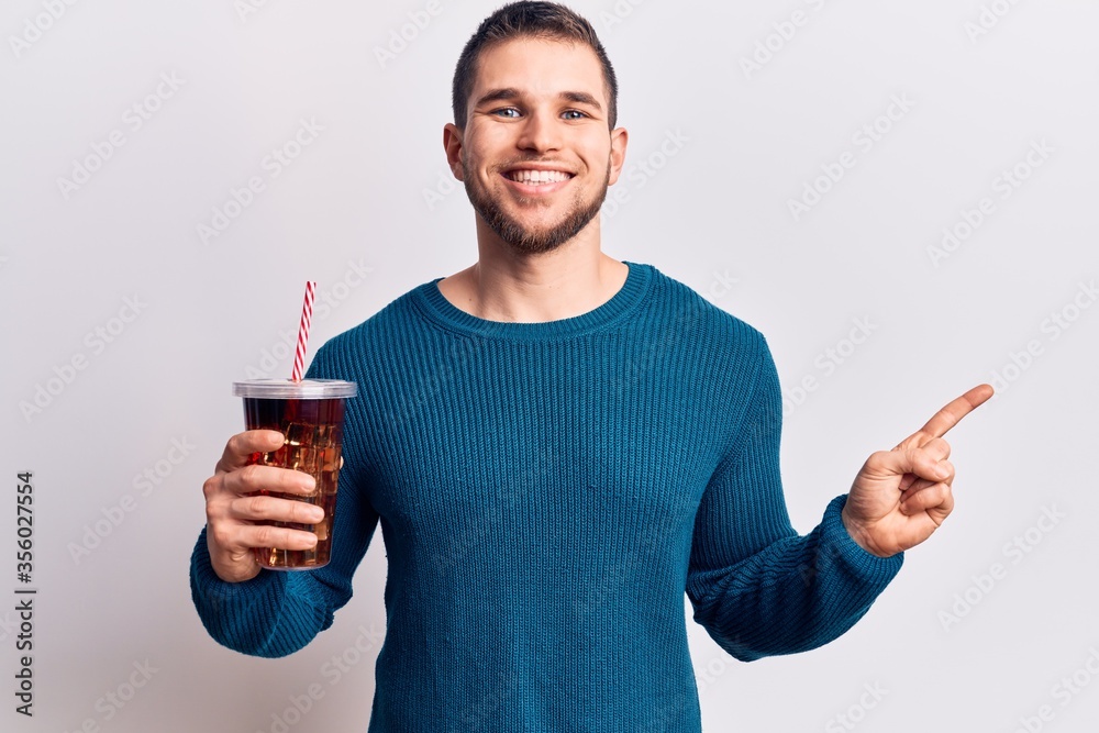 Young handsome man dirnking cola refreshment beverage smiling happy pointing with hand and finger to the side Young handsome man dirnking cola refreshment beverage smiling happy pointing with hand and finger to the side