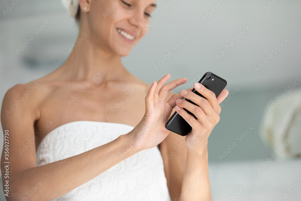 Close up smiling young woman with white bath towel on head using smartphone in bathroom, looking at screen, typing, chatting in social network, texting online after morning shower, reading news Close up smiling young woman with white bath towel on head using smartphone in bathroom, looking at screen, typing, chatting in social network, texting online after morning shower, reading news