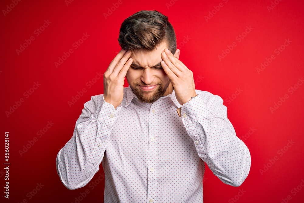 Young blond businessman with beard and blue eyes wearing elegant shirt over red background with hand on head for pain in head because stress. Suffering migraine. Young blond businessman with beard and blue eyes wearing elegant shirt over red background with hand on head for pain in head because stress. Suffering migraine.