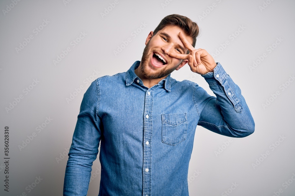 Young handsome blond man with beard and blue eyes wearing casual denim shirt Doing peace symbol with fingers over face, smiling cheerful showing victory Young handsome blond man with beard and blue eyes wearing casual denim shirt Doing peace symbol with fingers over face, smiling cheerful showing victory