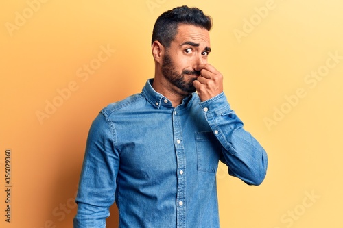 Young hispanic man wearing casual clothes smelling something stinky and disgusting, intolerable smell, holding breath with fingers on nose. bad smell