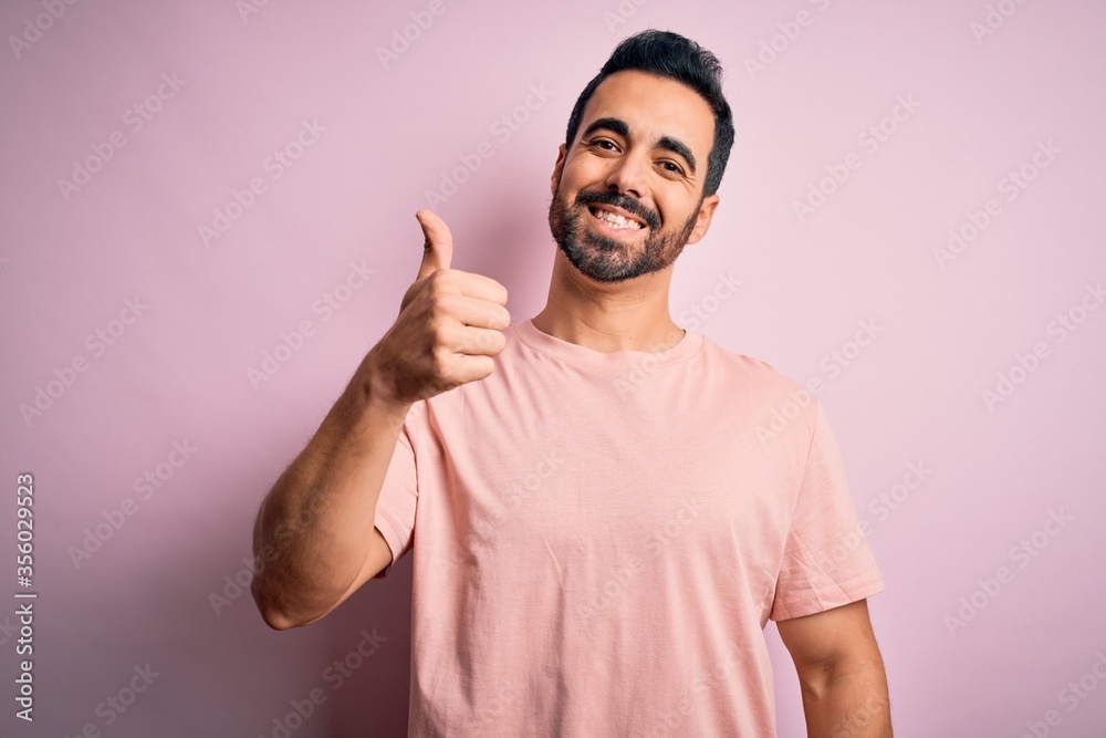 Young handsome man with beard wearing casual t-shirt standing over pink background doing happy thumbs up gesture with hand. Approving expression looking at the camera showing success. Young handsome man with beard wearing casual t-shirt standing over pink background doing happy thumbs up gesture with hand. Approving expression looking at the camera showing success.