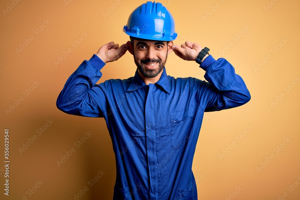 Mechanic man with beard wearing blue uniform and safety helmet over yellow background Smiling pulling ears with fingers, funny gesture. Audition problem