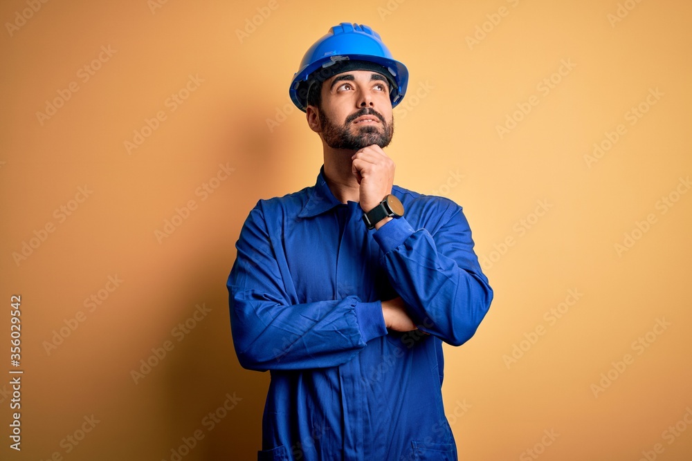 Mechanic man with beard wearing blue uniform and safety helmet over yellow background with hand on chin thinking about question, pensive expression. Smiling and thoughtful face. Doubt concept. Mechanic man with beard wearing blue uniform and safety helmet over yellow background with hand on chin thinking about question, pensive expression. Smiling and thoughtful face. Doubt concept.