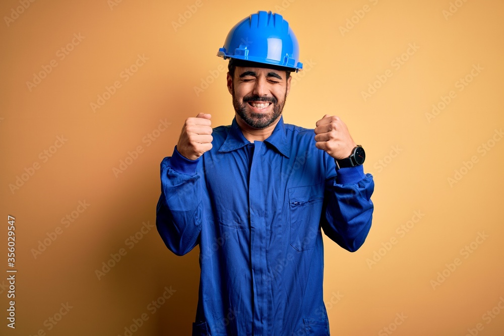 Mechanic man with beard wearing blue uniform and safety helmet over yellow background excited for success with arms raised and eyes closed celebrating victory smiling. Winner concept.