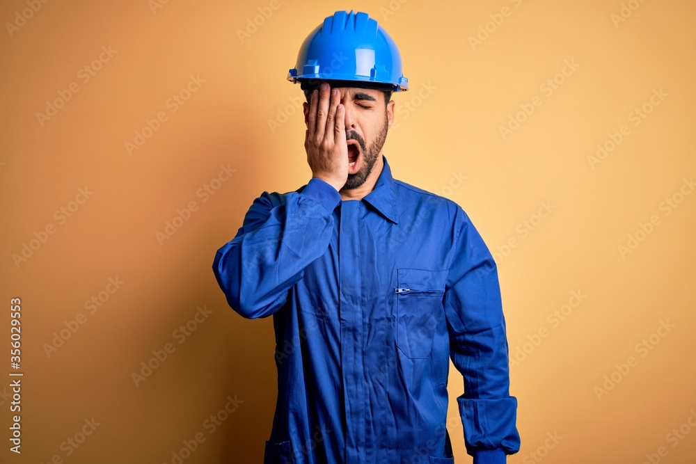 Mechanic man with beard wearing blue uniform and safety helmet over yellow background Yawning tired covering half face, eye and mouth with hand. Face hurts in pain.