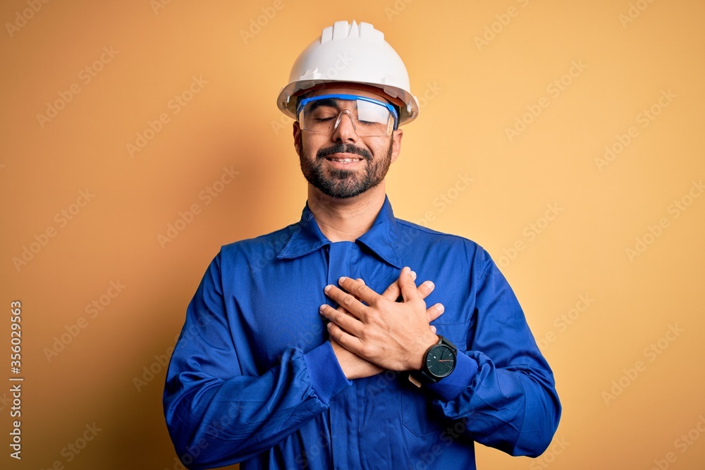 Mechanic man with beard wearing blue uniform and safety glasses over yellow background smiling with hands on chest with closed eyes and grateful gesture on face. Health concept.