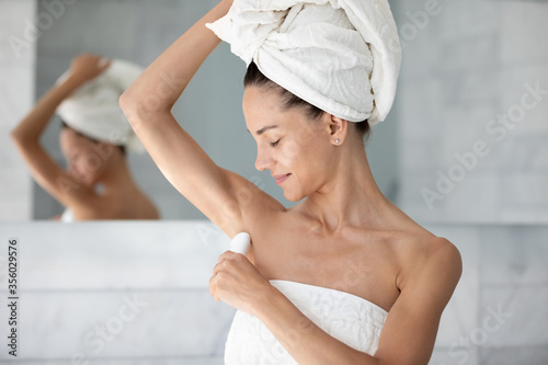 Satisfied young woman with white bath towel on head applying antiperspirant on armpit after shower, standing in modern bathroom, beautiful girl using stick underarm deodorant, morning routine