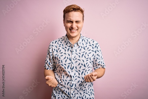 Young handsome redhead man wearing casual summer shirt standing over pink background very happy and excited doing winner gesture with arms raised, smiling and screaming for success. Celebration