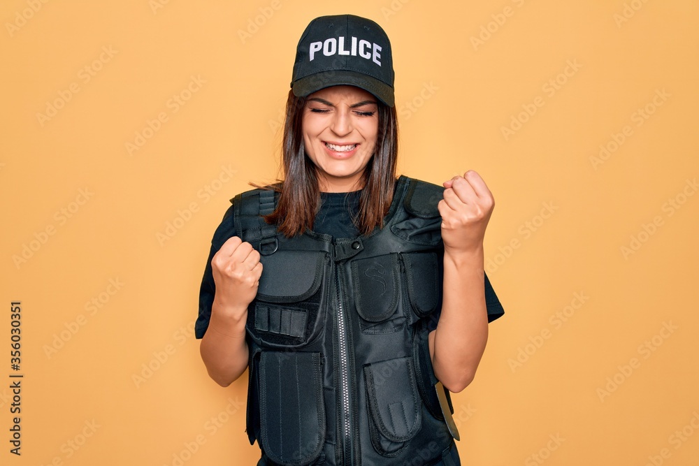Young beautiful brunette policewoman wearing police uniform bulletproof and cap celebrating surprised and amazed for success with arms raised and eyes closed. Winner concept. Young beautiful brunette policewoman wearing police uniform bulletproof and cap celebrating surprised and amazed for success with arms raised and eyes closed. Winner concept.
