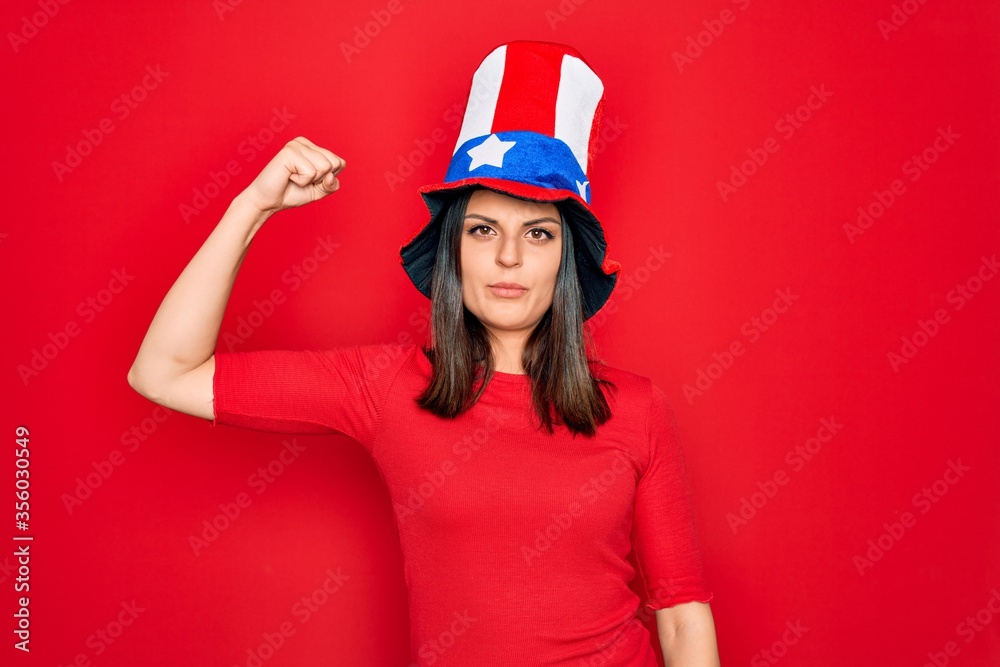 Young beautiful brunette woman wearing united states hat celebrating independence day Strong person showing arm muscle, confident and proud of power Young beautiful brunette woman wearing united states hat celebrating independence day Strong person showing arm muscle, confident and proud of power