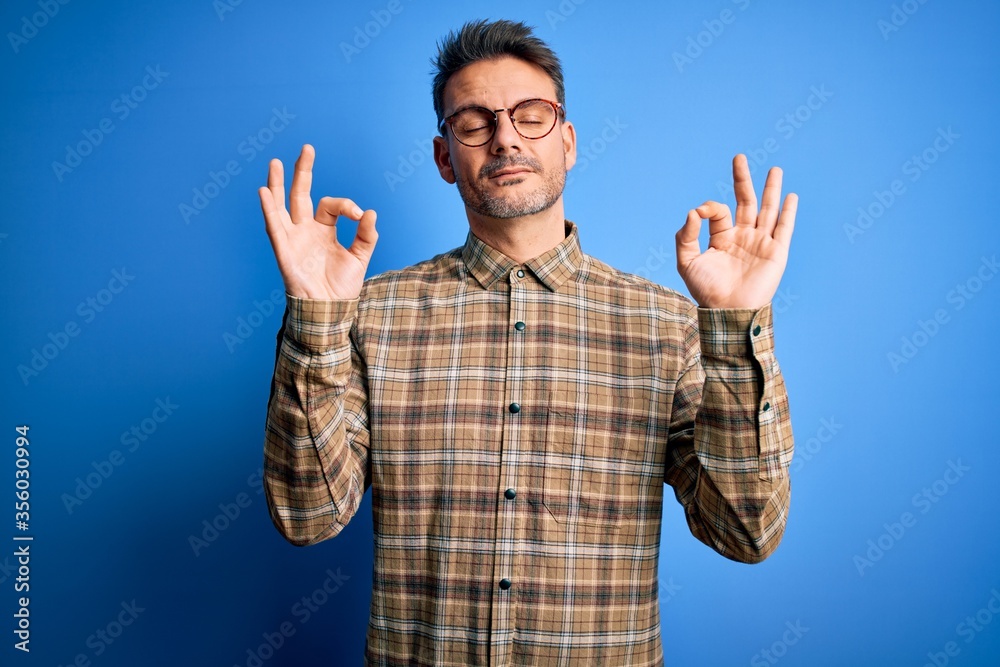 Naklejka premium Young handsome man wearing casual shirt and glasses standing over blue background relax and smiling with eyes closed doing meditation gesture with fingers. Yoga concept.