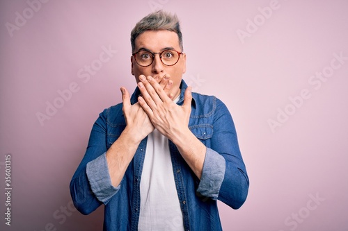 Young handsome modern man wearing glasses and denim jacket over pink isolated background shocked covering mouth with hands for mistake. Secret concept.