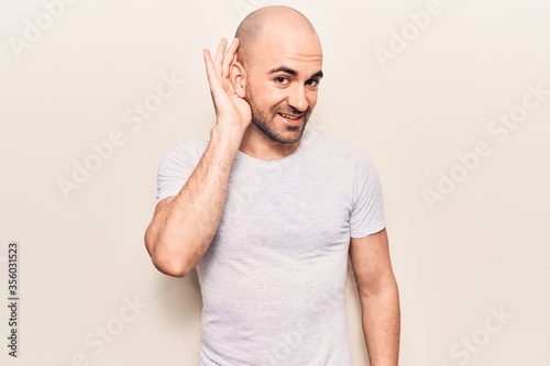 Young handsome bald man wearing casual t shirt smiling with hand over ear listening and hearing to rumor or gossip. deafness concept.