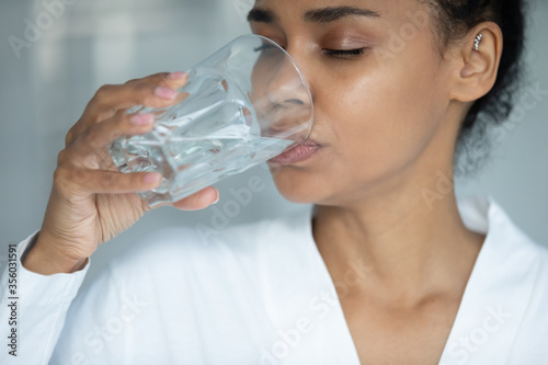 Close up thirsty beautiful African American young woman with perfect smooth skin drinking pure still mineral water, attractive girl holding glass, healthy lifestyle and habit concept, natural beauty
