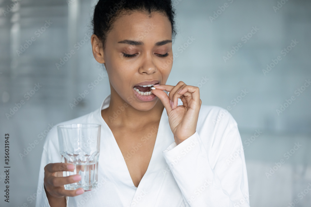 Close up smiling African American young woman taking round white pill, holding water glass, attractive girl wearing bathrobe taking supplements or vitamins in morning, healthcare treatment concept Close up smiling African American young woman taking round white pill, holding water glass, attractive girl wearing bathrobe taking supplements or vitamins in morning, healthcare treatment concept