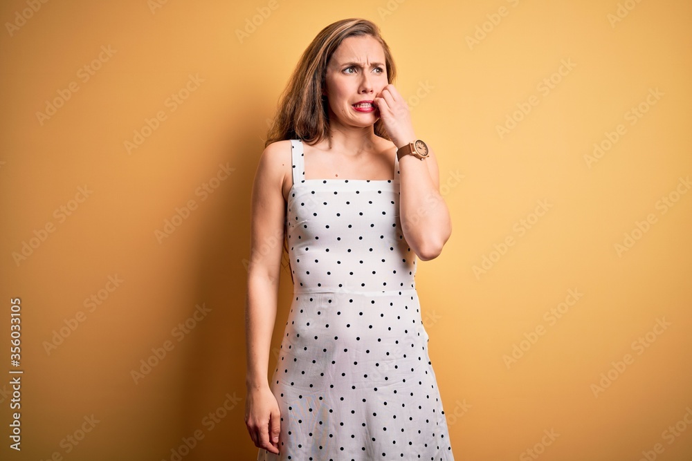 Young beautiful blonde woman on vacation wearing summer dress over yellow background looking stressed and nervous with hands on mouth biting nails. Anxiety problem. Young beautiful blonde woman on vacation wearing summer dress over yellow background looking stressed and nervous with hands on mouth biting nails. Anxiety problem.