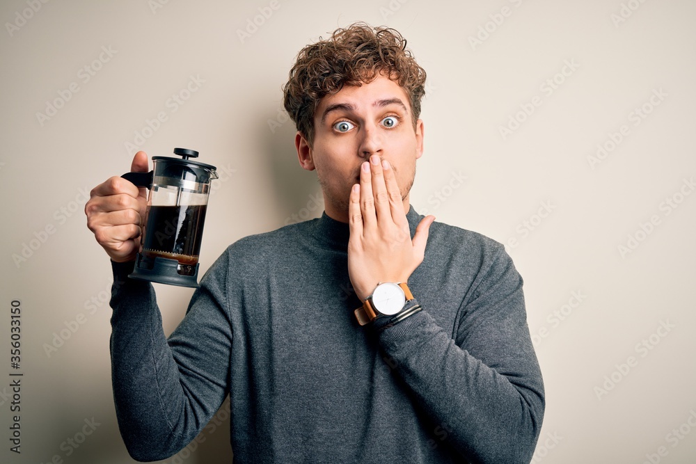 Young blond man with curly hair making coffee using coffemaker over white background cover mouth with hand shocked with shame for mistake, expression of fear, scared in silence, secret concept Young blond man with curly hair making coffee using coffemaker over white background cover mouth with hand shocked with shame for mistake, expression of fear, scared in silence, secret concept