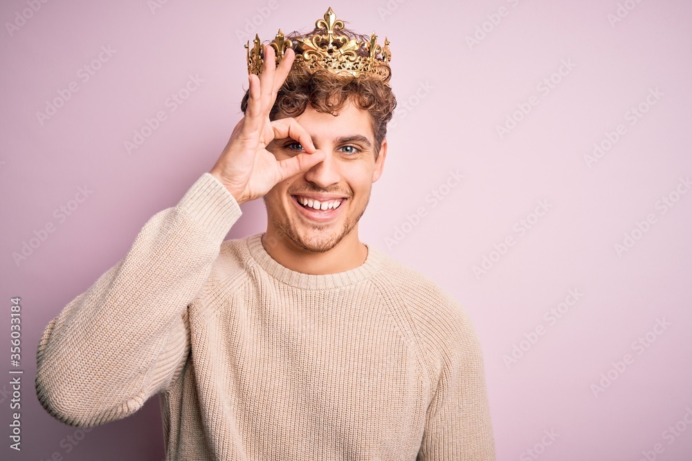 Φωτογραφία Young blond man with curly hair wearing golden crown of king over pink background doing ok gesture with hand smiling, eye looking through fingers with happy face