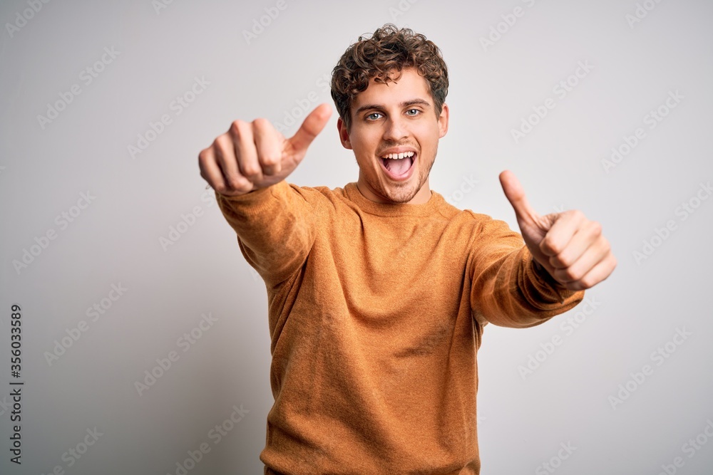 Young blond handsome man with curly hair wearing casual sweater over white background approving doing positive gesture with hand, thumbs up smiling and happy for success. Winner gesture. Young blond handsome man with curly hair wearing casual sweater over white background approving doing positive gesture with hand, thumbs up smiling and happy for success. Winner gesture.