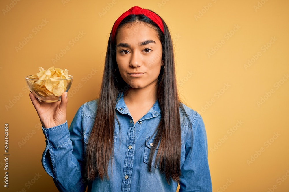 Young beautiful asian woman holding bowl with chips potatoes over isolated yellow background with a confident expression on smart face thinking serious Young beautiful asian woman holding bowl with chips potatoes over isolated yellow background with a confident expression on smart face thinking serious